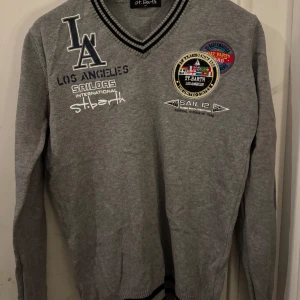 Grå v-ringad tröja St. Barth  - Grå v-ringad tröja från St. Barth med broderade patches och tryck på bröstet. Snygga svarta och vita ränder i kragen. Långärmad modell med sportig vibe och coola detaljer som LA-logga bland annat.