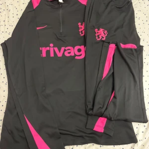 Chelsea träningsset Nike svart/rosa - Chelsea FC träningsset från Nike med långärmad tröja och byxor i svart med starka rosa detaljer. Tröjan har halv dragkedja, klubbmärke och Nike-logga på bröstet samt rosa axel- och ärmpaneler. Materialet är lätt och andas, perfekt för träning.