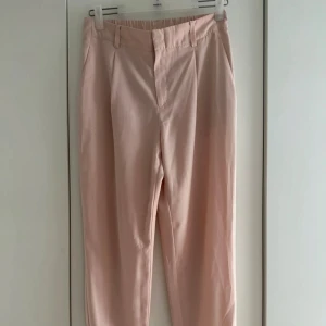 Ljusrosa byxor - Snygga ljusrosa byxor med resår bak. Byxorna har hällor för bälte, diskreta fickor bak och stängs med dragkedja och knapp framtill. Perfekta för en clean och fräsch look