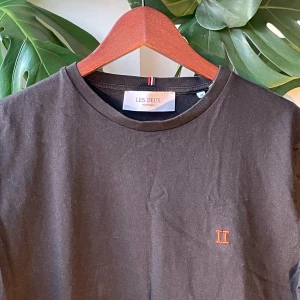 Svart t-shirt från Les Deux - Svart t-shirt från Les Deux med enkel design och orange broderad II-logga på bröstet. Klassisk rund halsringning och korta ärmar. Tillverkad i mjuk bomull som känns skön mot huden. Perfekt för en clean och stilren look.