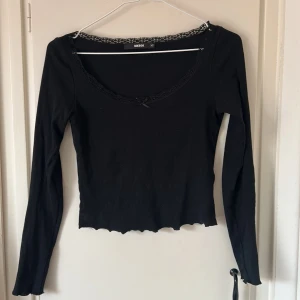 Svart topp med spets från BikBok - Supersöt svart långärmad topp från BikBok i ribbad bomull. Urringad hals med spetskant och liten rosett framtill. Croppad modell med vågig nederkant, perfekt att styla med jeans eller kjol.