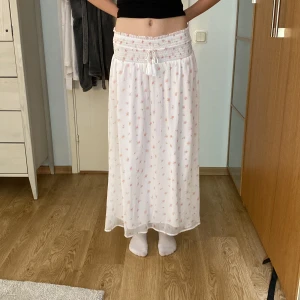 Vit blommig långkjol från Gina Tricot - Supersöt långkjol från Gina Tricot i vitt med små rosa blommor. Kjolen har smockad midja med snörning och är gjord i ett lätt, luftigt tyg som känns perfekt för varma dagar. Den har en flowy passform och romantisk vibe, perfekt att styla med crop top. Använd ca 2 gånger.