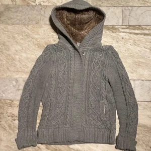 Grå stickad cardigan - Mysig grå stickad cardigan med kabelstickat mönster och mjukt foder i luvan. Perfekt för kyliga dagar med sin varma och bekväma design. Långa ärmar och ribbade muddar ger en skön passform.