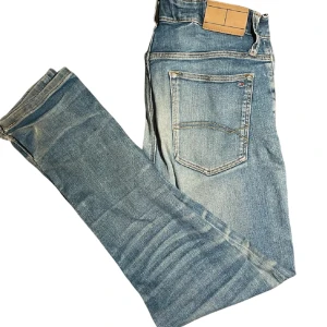 Ljusblå Tommy Hilfiger jeans med raka ben - Säljer ett par ljusblå Tommy Hilfiger jeans med klassisk femficksdesign och raka ben. Jeansen har slitningar och tvättade detaljer för en avslappnad look. Passar perfekt till vardags och har en neutral läderpatch bak i midjan.
