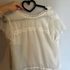 Vit prickig blus med volangärm - Superfin vit blus i transparent tyg med små prickar och volangärmar. Blusen har rund halsringning med rynkade detaljer och spetsband över bröstet. Perfekt för en romantisk look och passar till både jeans och kjol.