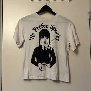 Vit Wednesday t-shirt från H&M - Vit t-shirt från H&M med tryck av Wednesday Addams och texten 'We Prefer Spooky' på framsidan. Klassisk rund hals och korta ärmar. Materialet är mjukt och skönt, perfekt för dig som gillar en cool och unik stil.