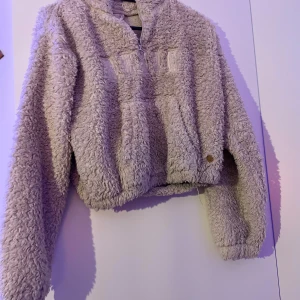 Beige teddyjacka med half zip - Supermysig beige teddyjacka från H&M med stor ring i dragkedjan och broderad text framtill. Jackan har lång ärm, hög krage och är tillverkad i fluffigt teddy-material. Perfekt för kalla dagar när du vill vara både varm och snygg.