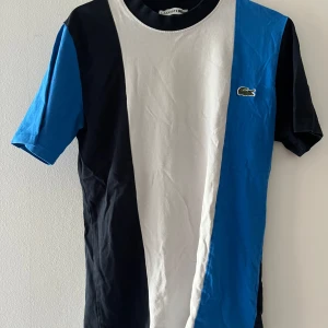 Lacoste t-shirt med blockfärger - Snygg t-shirt från Lacoste med breda vertikala block i blått, vitt och svart. Klassisk krokodillogga på bröstet. Skön passform och rund halsringning, perfekt för en stilren och sportig look.
