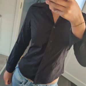 Svart skjorta från Sisters, stl 34 - Klassisk svart skjorta från Sisters i storlek 34. Skjortan har lång ärm, knappar framtill och en figurnära passform. Perfekt till jeans eller kjol för en clean look. Materialet känns lätt och bekvämt mot huden.