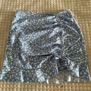 Blå blommig kortkjol med volang - Supersöt blå kortkjol med vita blommor och volangkant. Kjolen har snörning och rynkdetalj framtill som ger en snygg form. Perfekt för dig som gillar en feminin och trendig stil. Dragkedja i sidan för enkel påklädning.