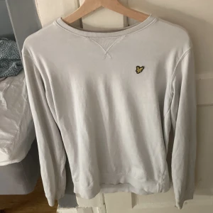 Vit sweatshirt från Lyle & Scott - Vit sweatshirt från Lyle & Scott med klassisk rund hals och broderad gul logga på bröstet. Tröjan har långa ärmar och ribbade muddar vid ärmslut och nederkant. Perfekt för en clean och stilren look.