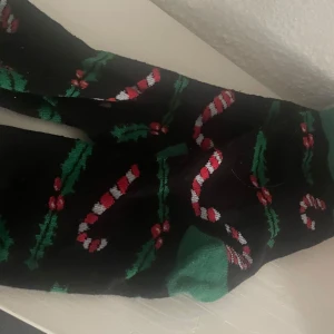 Svarta julstrumpor med polkagrisar - Svarta strumpor med gröna tår och hälar, dekorerade med röda och vita polkagrisar samt gröna julblad. Perfekta för att maxa julkänslan och sticka ut med ett festligt mönster. Mjuka och sköna, passar till alla dina vinterlooks.