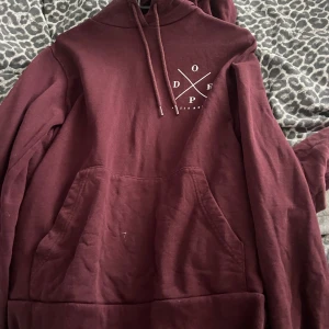 Vinröd hoodie från Dope - Säljer en vinröd hoodie från Dope med vit logga tryckt både fram och bak. Tröjan har huva med snörning och en stor magficka. 
