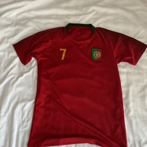 Portugal Ronaldo 7 fotbollströja röd - Snygg röd Portugal fotbollströja med gröna detaljer i kragen och guldiga tryck. Nummer 7 och Ronaldo på ryggen, samt landslagets emblem på bröstet. Perfekt för dig som vill hylla Cristiano Ronaldo. Tillverkad i lätt och ventilerande material. 