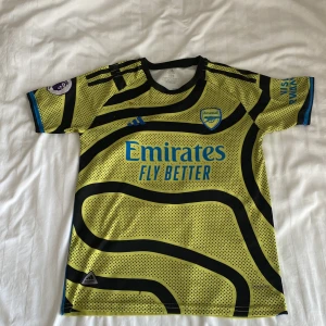 Arsenal bortatröja G.Jesus #9 Adidas - Arsenal bortatröja med G.Jesus och nummer 9 på ryggen. Tröjan är gul med svarta vågiga linjer och prickar, blå detaljer och klubbmärke på bröstet. Tillverkad i lätt och ventilerande polyester, kortärmad modell. Adidas-logga och sponsortryck i blått.