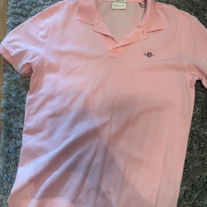 Rosa pikétröja från GANT - Säljer en klassisk rosa pikétröja från GANT med broderad logga på bröstet. Tröjan har korta ärmar, krage och knäppning vid halsen. Perfekt för en fräsch och stilren look. Tillverkad i mjuk bomull som andas.