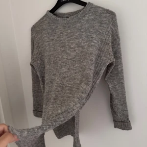 Grå stickad tröja med rund hals - Mysig grå stickad tröja med rund hals och långa ärmar. Tröjan har ribbade muddar vid ärmslut och nederkant, vilket ger en avslappnad och skön känsla. Perfekt att slänga på sig när det är kyligt ute.