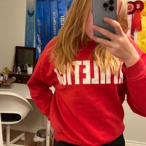 Röd sweatshirt med vit text - Röd sweatshirt med stor vit text 'ATHLETIC' framtill. Klassisk rund halsringning och ribbade muddar vid ärmslut och nederkant. Perfekt för en sportig och avslappnad stil.