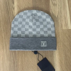 Grå rutig mössa från Louis Vuitton - Snygg grå mössa från Louis Vuitton med klassiskt rutigt mönster i ljusgrått och vitt. Mössan har en bred uppvikt kant med LV-logga framtill. Tillverkad i mjukt stickat material som känns lyxigt och passar perfekt till kalla dagar.
