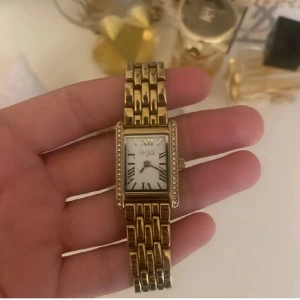 Guldig Regal klocka med stenar - Snygg guldig klocka från Regal med rektangulärt ur och romerska siffror på vit urtavla. Länken är i metall och boetten är dekorerad med små gnistrande stenar längs kanterna. Perfekt accessoar för att lyfta din stil.