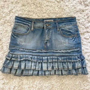 Jeanskjol med volanger från Cindy M - Supersöt blå jeanskjol från Cindy M med volangkant och slitna detaljer. Kjolen har klassiska fickor fram och bak, bälteshällor och stängs med knapp och dragkedja. Materialet är stretchigt med 98% bomull och 2% elastan för en bekväm passform.