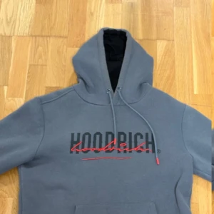 Hoodrich hoddie grå  - Snygg grå hoodie från Hoodrich med svart och rött tryck på både fram- och baksidan. Tröjan har huva med snörning och en klassisk känguruficka framtill. 