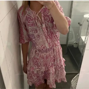 Rosa och vit kortklänning med volang - Supersöt kortklänning i rosa och vitt med paisleyinspirerat mönster. Klänningen har volangdetaljer, knytning vid halsen och halvlånga ärmar