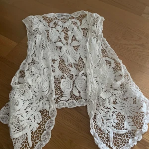 Vit spetsväst från ONLY Love Collection - Superfin vit väst från ONLY Love Collection i spets med blommiga detaljer. Västen är öppen framtill och har ett luftigt, bohemiskt mönster som gör den perfekt att slänga över en topp eller klänning. Snygg och unik detalj till din outfit!