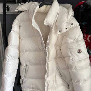 Moncler Maya - Vit - Säljer en snygg vit pufferjacka från Moncler med huva och dragkedja. Jackan har två fickor med dragkedja framtill och en ärmficka med Moncler-logga. Perfekt för kalla dagar och riktigt stilren look. Köpt 2022, använd
