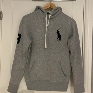Grå hoodie från Polo Ralph Lauren - Säljer en grå hoodie från Polo Ralph Lauren med stor svart logga på bröstet och siffra på ärmen. Tröjan har huva med snörning, känguruficka och är tillverkad i mjuk bomullsmix. Perfekt för chill dagar eller när du vill ha en clean streetstyle.