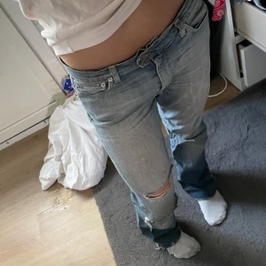 Ljusblå slitna jeans med hål - Säljer ett par ljusblå jeans med slitningar och ett stort hål på ena knät. Jeansen har låg midja och klassisk femficksmodell. De är raka i modellen och har en avslappnad vibe, perfekta för dig som gillar en edgy streetstil.