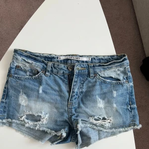 Blå slitna lågmidjadd jeansshorts Fabulous S - Snygga blå jeansshorts från Fabulous i storlek S. De har slitna detaljer, fransig kant och klassisk femficksmodell. Perfekta för sommaren med sin avslappnade och trendiga look. Passar dig som gillar en lite tuffare stil.