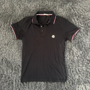 Svart Moncler pikétröja med logga - Svart pikétröja från Moncler med klassisk krage och knäppning framtill. Röd, vit och blå rand på krage och ärmslut ger en sportig vibe. Broderad Moncler-logga på bröstet. Tillverkad i bomull för skön känsla och snygg passform.