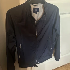 Mörkblå vindjacka från Gant, M - Snygg mörkblå vindjacka från Gant med dragkedja framtill och två fickor med dragkedja. Jackan har elastisk nederkant. Perfekt för dig som gillar stilren och sportig look. Storlek M men sitter bättre som storlek S. Nypris 1500kr