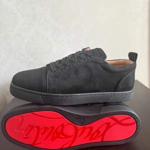 Christian Louboutin svart  - Svarta sneakers i mocka från Christian Louboutin med ikonisk röd sula och diskret logga undertill. Klassisk låg modell med rund tå, svarta snören och snygga detaljer. Insidan är ljusbeige och skon har en stilren, lyxig vibe. 42 men passar 41 . Använda 1-2 gånger bara men syns knappt. 