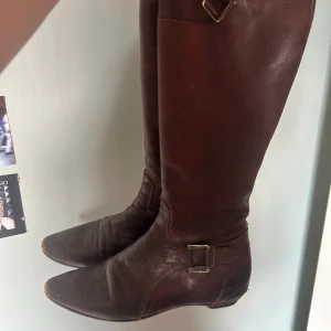 Bruna höga skinnstövlar med spänne - Säljer ett par bruna höga stövlar i skinn med snyggt spänne vid ankeln. De har spetsig tå och platt sula, perfekt till kjol eller klänning. Klassisk modell som funkar till många olika stilar.