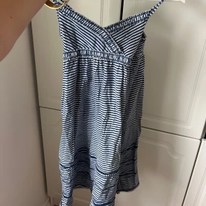 Randig blåvit sommarklänning - Söt kortklänning med smala axelband och omlott vid bysten. Klänningen är blå- och vitrandig med resår i midjan och lätt utställd kjol. Perfekt för varma dagar och har en avslappnad, luftig känsla. Materialet känns mjukt och bekvämt.