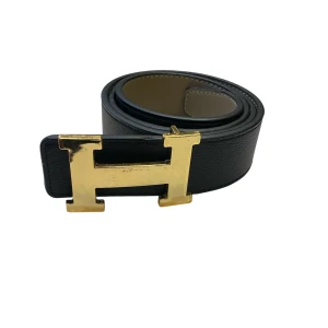 Svar skärp med guldfärgat spänne  - Snyggt svart skärp i äkta skinn med spänne i guldmetall. Bältet har en klassisk och stilren design som verkligen sticker ut med sitt stora, blanka spänne. Perfekt för att lyfta vilken outfit som helst.