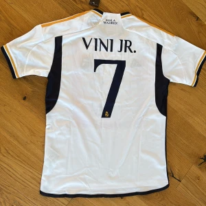 Real Madrid Vini Jr. 7 Adidas tröja - Snygg Real Madrid matchtröja med Vini Jr. och nummer 7 på ryggen. Vit bas med marinblå detaljer och guldiga ränder på axlarna. Broderat klubbmärke och Adidas-logga framtill, samt 'Emirates Fly Better' tryck. Tillverkad i lätt och ventilerande polyester. Och aldrig använd 