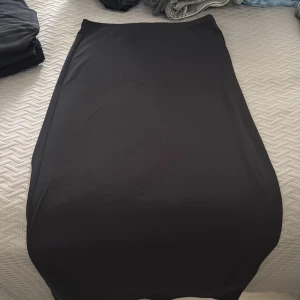 Svart långkjol i stretchigt tyg - Enkel och stilren svart långkjol med rak passform. Kjolen är gjord i ett satin-likt stretchigt material som sitter snyggt och bekvämt. Perfekt för dig som gillar minimalistisk stil och vill ha en kjol som funkar till många olika looks.