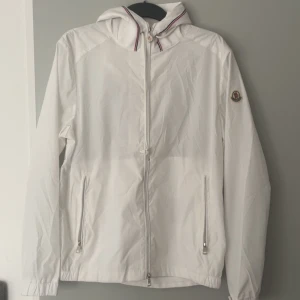 Moncler jacka - Snygg vit vindjacka från Moncler med huva och dragkedja framtill. Jackan har två fickor med dragkedja och Moncler-logga på ärmen. Lätt och stilren design, perfekt för vår och sommar. Materialet är polyester och jackan har långa ärmar.