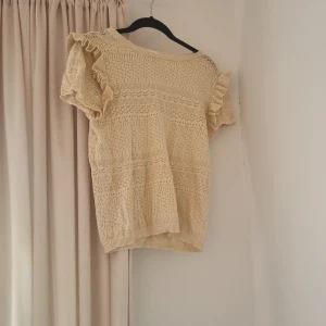 Beige stickad blus med volangärm XS - Supersöt beige stickad blus från Stories i storlek XS. Blusen har korta ärmar med volangdetaljer och ett vackert hålmönster över hela plagget. Perfekt för vår och sommar när du vill ha en romantisk och luftig look.