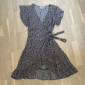 Blommig omlottklänning från Vila - Supersöt kort omlottklänning från Vila med svart botten och små blommor i lila, vitt och gult. Klänningen har volangkant, knyt i midjan, v-ringning och korta ärmar. Perfekt för sommarens alla tillfällen!