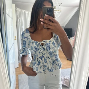 Blommig blus med volang och knyt - Supersöt blommig blus i vitt och blått med smockad midja, volangkant och korta puffärmar. Snygg knytning framtill och lite djup i  ryggen. Endast använd fåtal gånger, storlek S! 