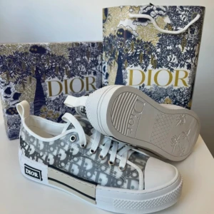 Dior sneakers med monogram i grått - Snygga Dior sneakers med ikoniskt grått och vitt monogrammönster. Skorna har vita snören, rund tå och platt sula med präglade detaljer undertill. Ovandelen är i syntetmaterial och har en modern, stilren look med tydlig Dior-logga på sidan.
