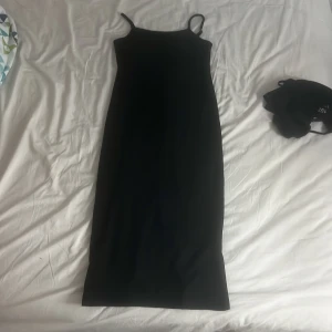 Svart långklänning med smala axelband - En stilren svart långklänning med smala axelband och rak passform. Perfekt för dig som gillar minimalistisk och tidlös stil. Klänningen är ärmlös och har en enkel, elegant siluett som passar till många olika tillfällen.