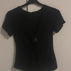 Svart omlottopp med dekorerad detalj - Svart omlottopp med kort ärm och djup v-ringning. Toppen har en snygg knytning framtill och en rund dekorerad detalj i mitten med pärlliknande mönster. Perfekt för dig som gillar stilrena plagg med en twist.