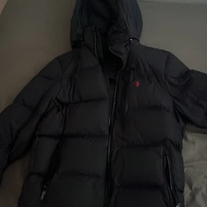 Svart pufferjacka från Polo Ralph Lauren - Säljer en svart pufferjacka från Polo Ralph Lauren med huva och dragkedja. Jackan har en klassisk quiltad look, två fickor med dragkedja och den ikoniska röda Polo-loggan på bröstet. Perfekt för kalla dagar när du vill hålla stilen.