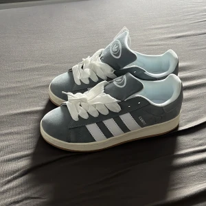 Adidas Campus grå sneakers - Grå Adidas Campus sneakers med klassiska vita ränder på sidorna och vit sula. Skorna har breda vita skosnören och tydlig Adidas-logga på plösen. Ovandelen är i mocka och insidan är vit. Perfekta för en stilren och sportig look.
