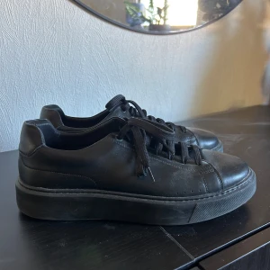 Svarta sneakers från Hugo Boss - Nypris 1500kr. Stilrena svarta sneakers från Hugo Boss med diskret röd logga på hälen och röd innersula. Skorna har en chunky platt sula, rund tå och är tillverkade i skinn. Klassisk snörning framtill och minimalistisk design som passar till det mesta.
